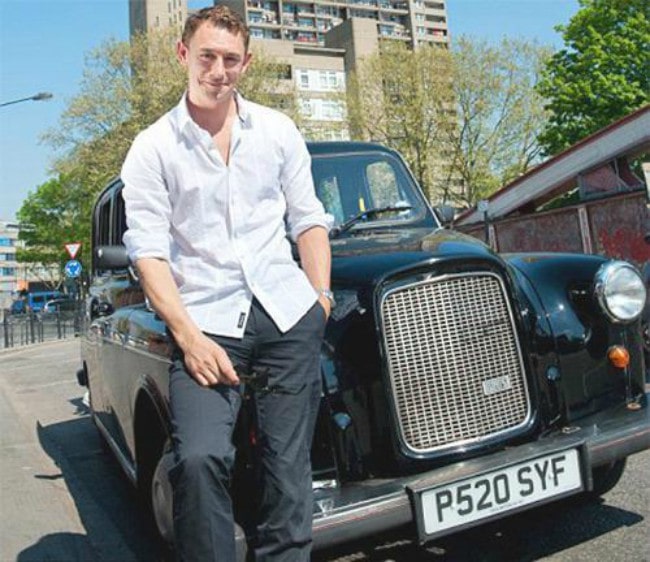 JJ Feild in a white shirt and black pants posing in front of his car.